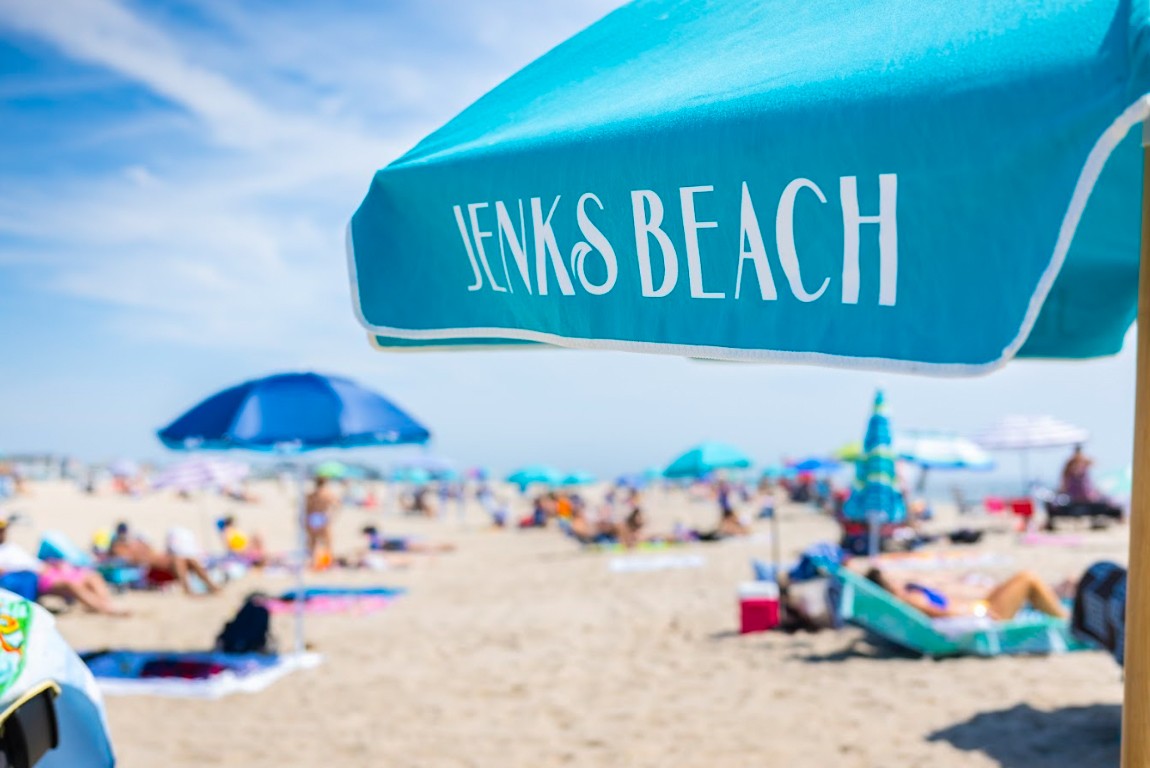 A turquoise beach umbrella labeled "Jenks Beach" with people relaxing on the sandy beach in Point Pleasant Beach, NJ.