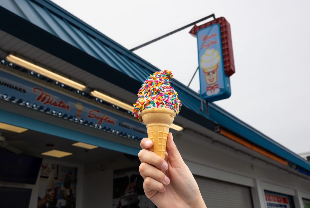 Hand holding a colorful, sprinkle-topped ice cream cone in front of a retro ice cream shop with a blue roof and "Mister Softee" signage at Jenkinson's Boardwalk in Point Pleasant Beach New Jersey